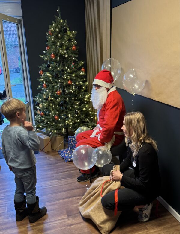 Weihnachtsmann vor festlich geschmücktem Baum mit leuchtenden Heliumballons, seine Helferin sitzt daneben, ein kleiner Junge steht vor ihm und zeigt begeistert nach oben.